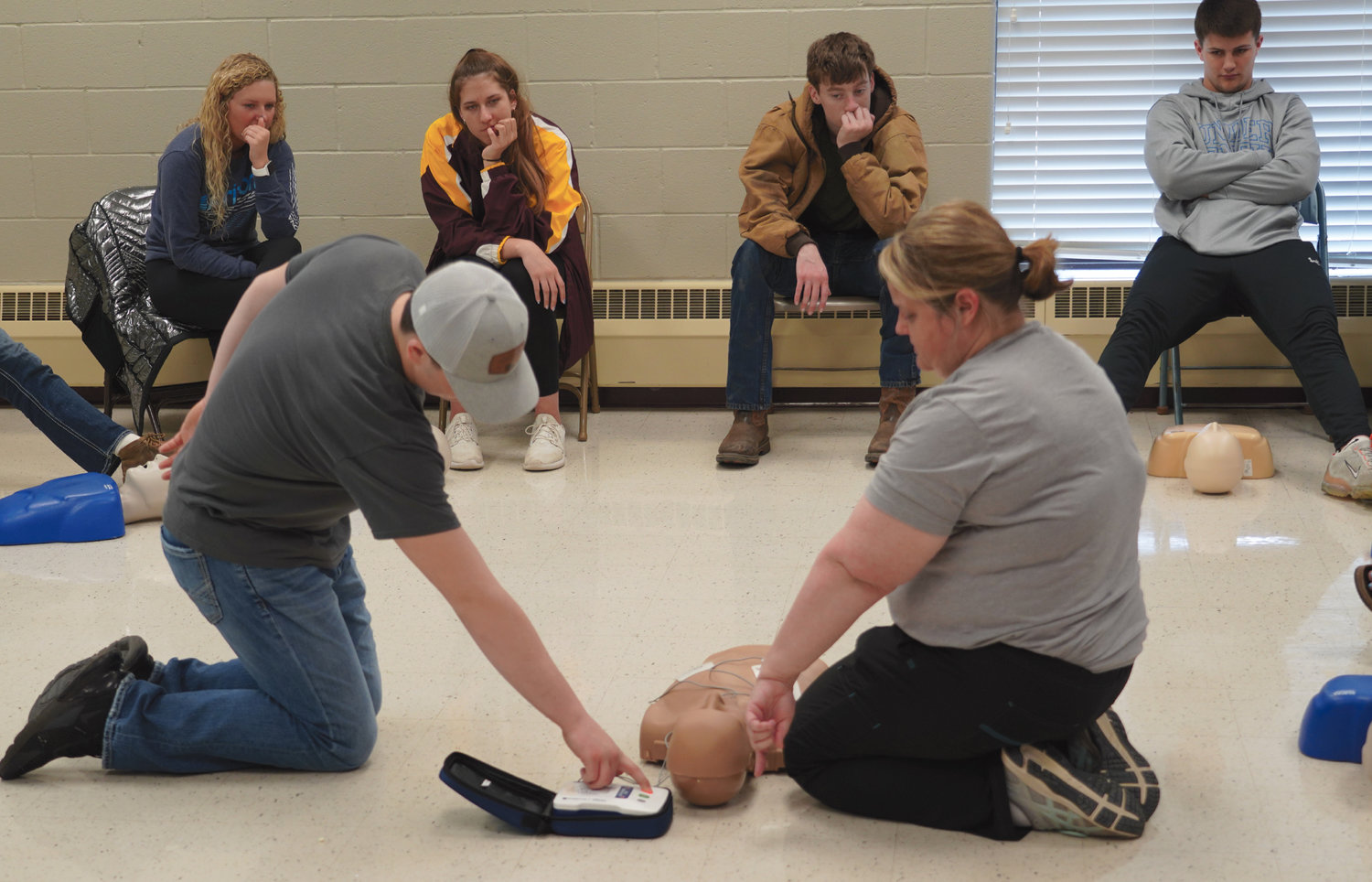 De Smet seniors are participating in a CPR class required by South Dakota before graduation. The class assembled in the community classroom of the armory on Wednesday and watched as Jodi Jung, CNP, right, who works for Horizon Health Care, demonstrates the use of an Automated External Defibrillator (AED), while Cody Aughenbaugh presses the button on the AED to deliver a lifesaving shock.